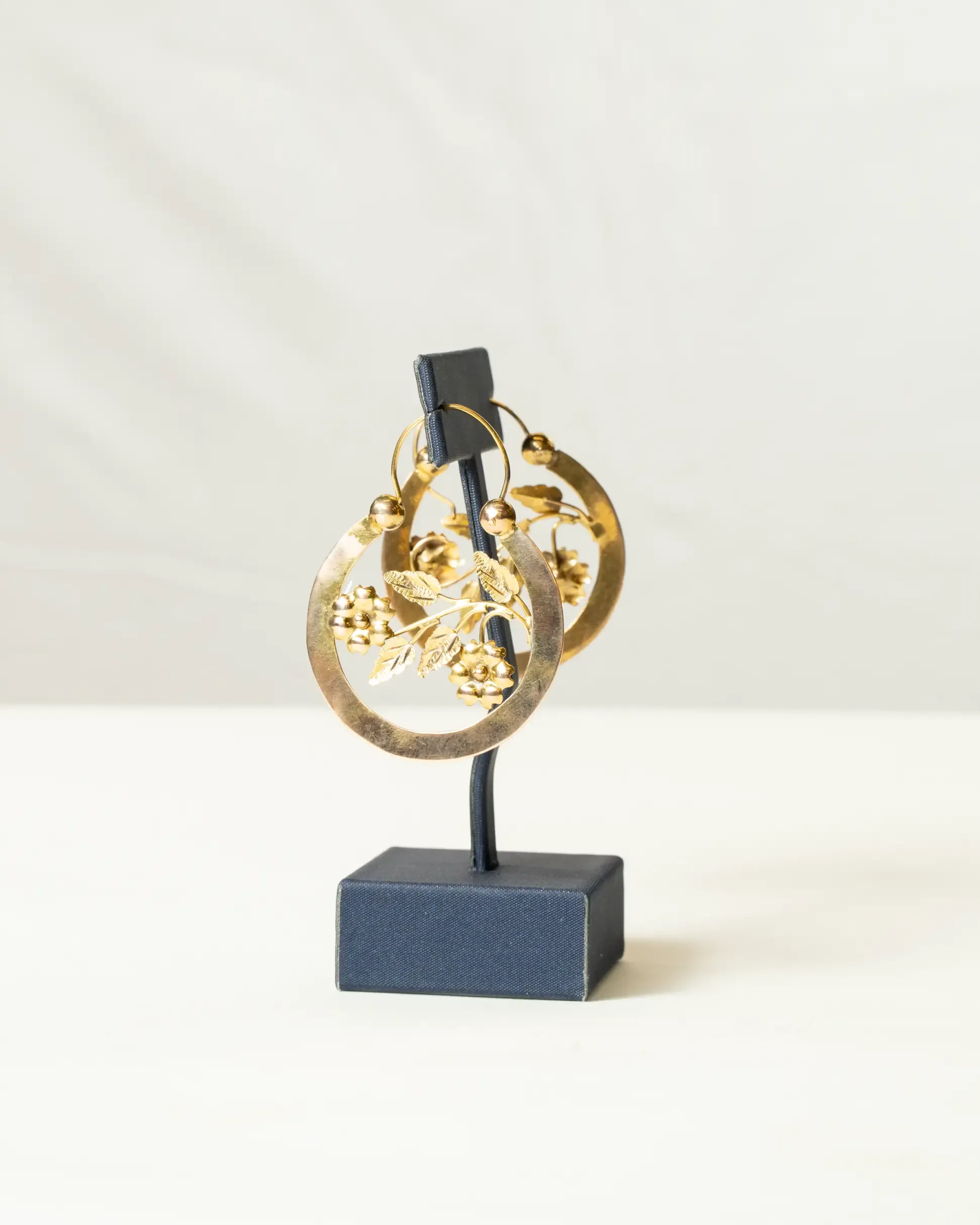 Gold hoop earrings with intricate floral and leaf detailing, displayed on a navy blue stand against a light background.