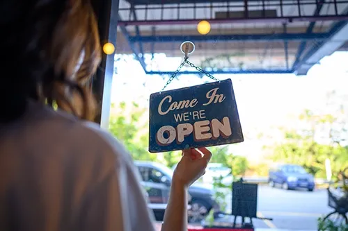 A person hanging a sign that reads "Come In, We're Open" on a glass door, indicating a business is open.